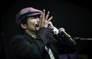 Bonny B spielt Mundharmonica auf der Bühne und trägt einen schwarzen Anzug und eine fraue Mütze