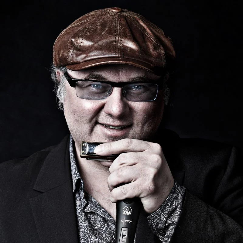 Dozent Chris Kramer trägt eine braune Lederkappe und hält in der Hand eine Mundharmonika und ein Mikrofon