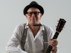 Dozent Eddie Martin er trägt ei weißes Hemd und einen Hut, hält eine Gitarre in der Hand