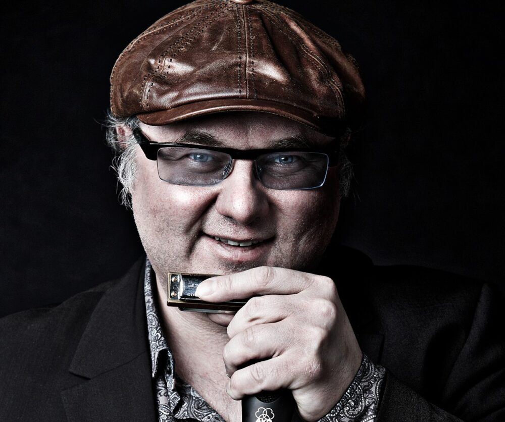 Dozent Chris Kramer trägt eine braune Lederkappe und hält in der Hand eine Mundharmonika und ein Mikrofon