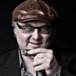 Dozent Chris Kramer trägt eine braune Lederkappe und hält in der Hand eine Mundharmonika und ein Mikrofon